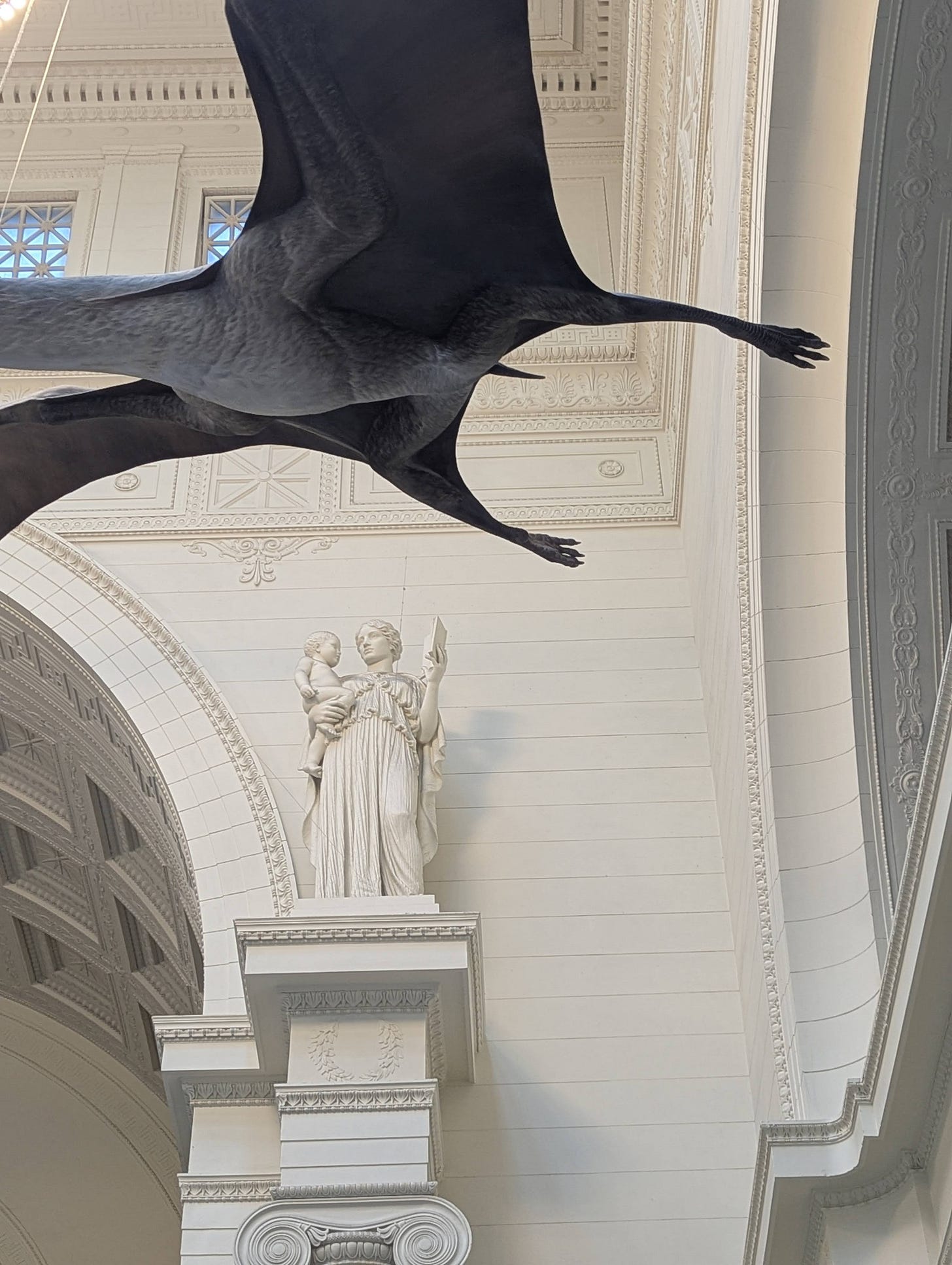 an image of a statue of a woman holding an open book in one hand and a toddler on the opposite hip. In the foreground, suspended above the statue, is an enormous pterodactyl model an image of a statue of a woman holding an open book in one hand and a toddler on the opposite hip. In the foreground, suspended above the statue, is an enormous pterodactyl model