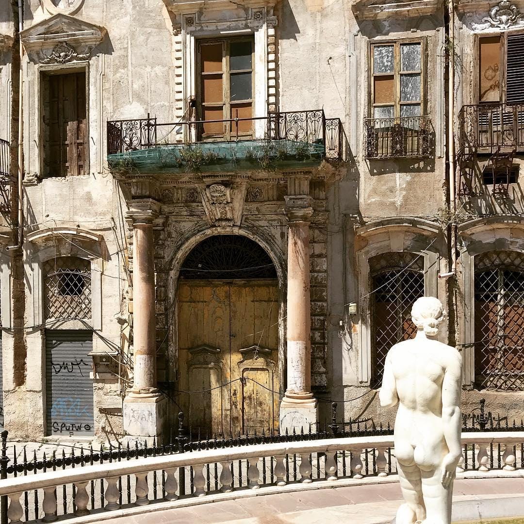 A life-sized sculpture in a fountain facing a decrepit building in Palermo, Sicily on a sunny day. Wrought iron covers the lower windows, the wooden front doors are flanked by sepia baroque pillars