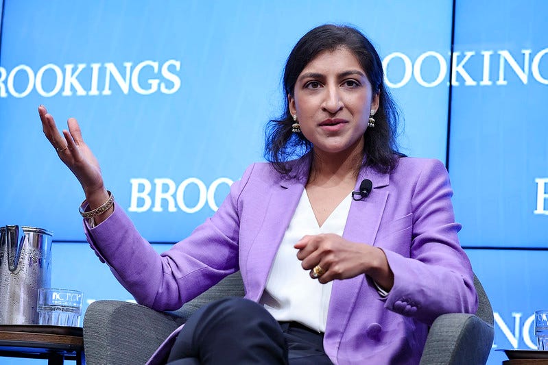 FTC chair Lina Khan, wearing a nifty purple blazer, gestures with her right hand while speaking during a panel discussion. Behind her, a wall of monitors displays the Brookings Institution's logo on a light blue background.