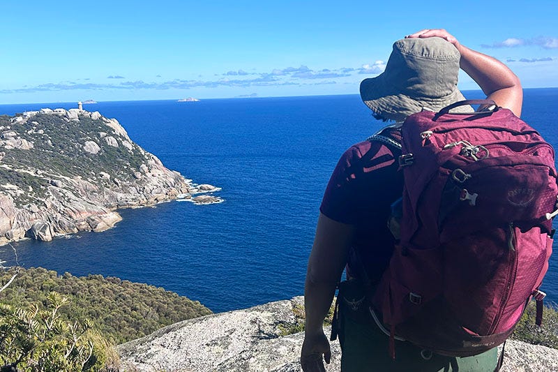 A woman in hiking gear with a backpack stands in the foreground, in the background is the ocean and a light station.