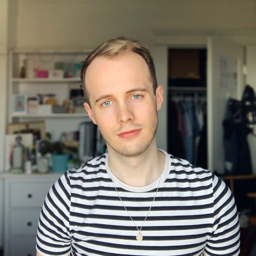 Photo of a white person with short blonde hair and blue eyes, wearing a black and white striped shirt, looking softly and directly into the lens.