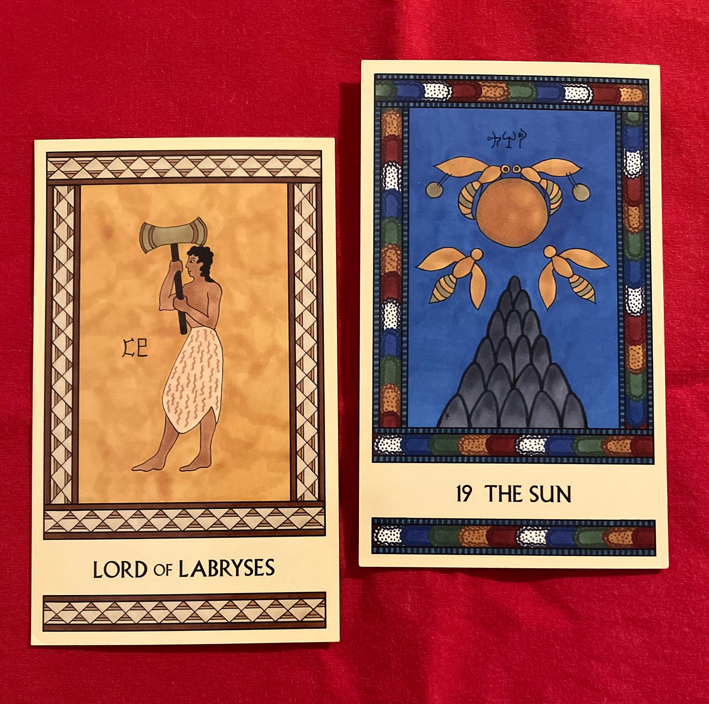 Two Minoan Tarot cards side by side. The Lord of Labryses is in shades of gold and cream. It shows a Minoan man wearing an animal hide skirt, facing left, holding a large labrys. The Sun has a multicolored border. It shows four bees flying near a deep golden Sun above a grey mountaintop, with a deep blue sky surrounding them.