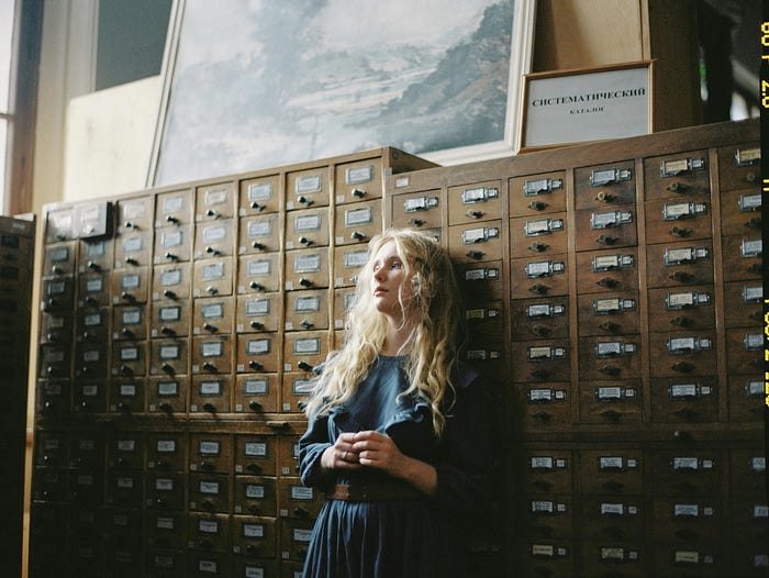 A woman in a blue dress leaning back against an archived stack of documents, looking a little sad.