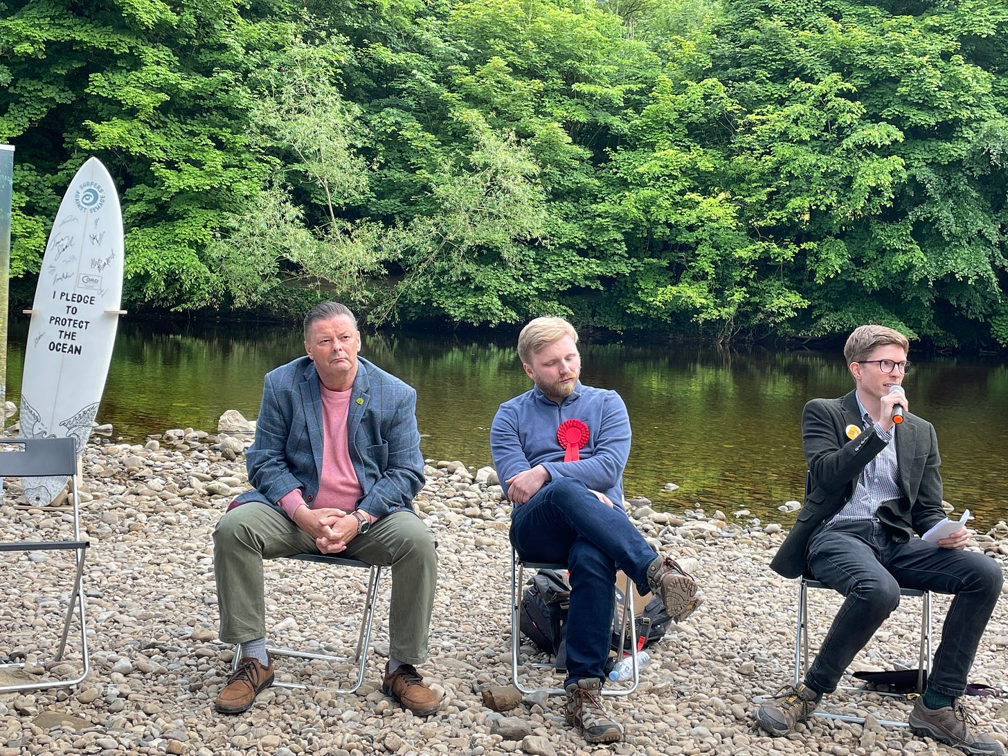 Election candidates sit on temporary chairs by the river swale Kevin Foster (Green), Tom Wilson (Labour) and Daniel Callaghan (Lib Dem)