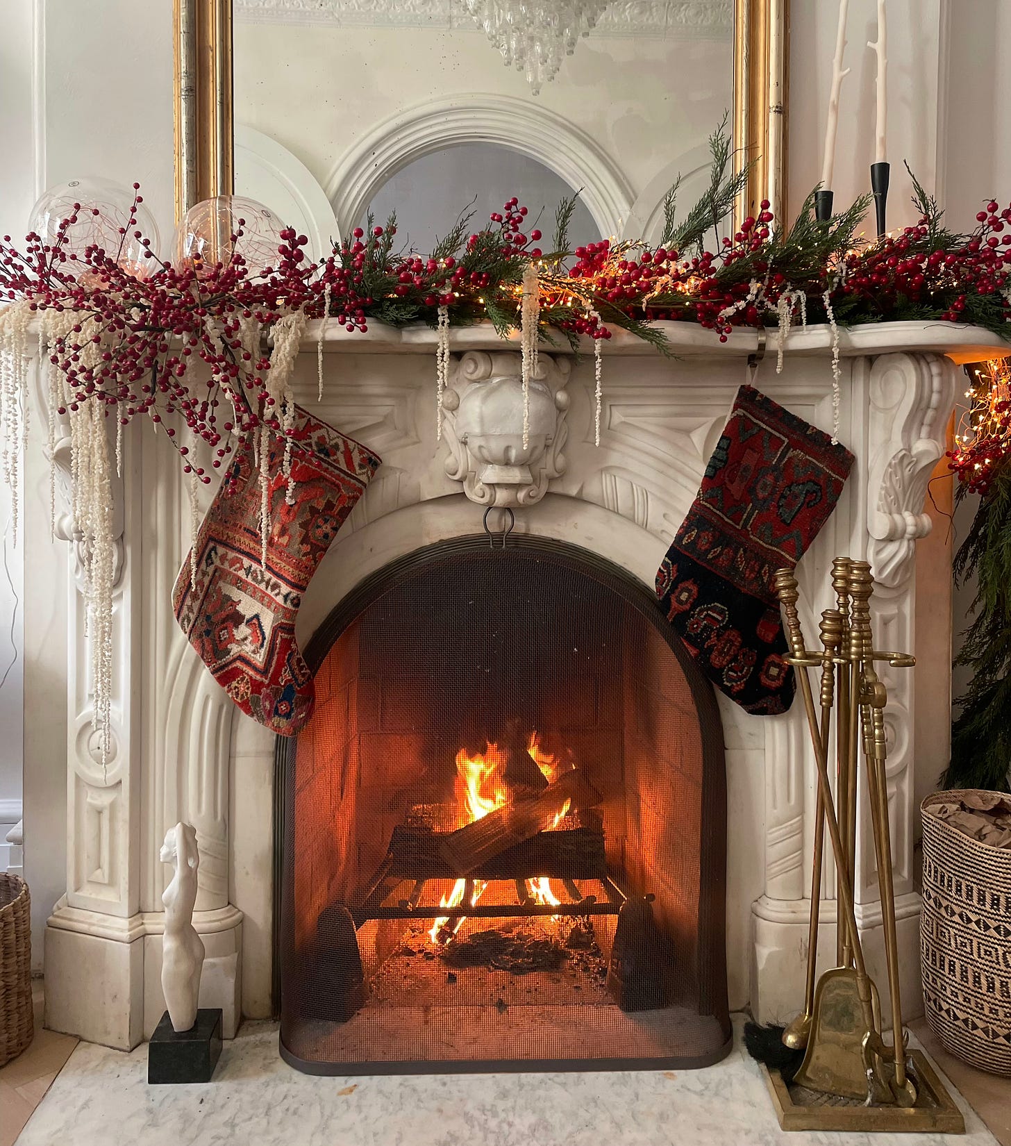 An ornate marble fireplace is decorated for Christmas: red berries, pine boughs, pearls and twinkle lights adorn the mantle, along with two large stockings made from North African textiles. In the fireplace, a fire glows.
