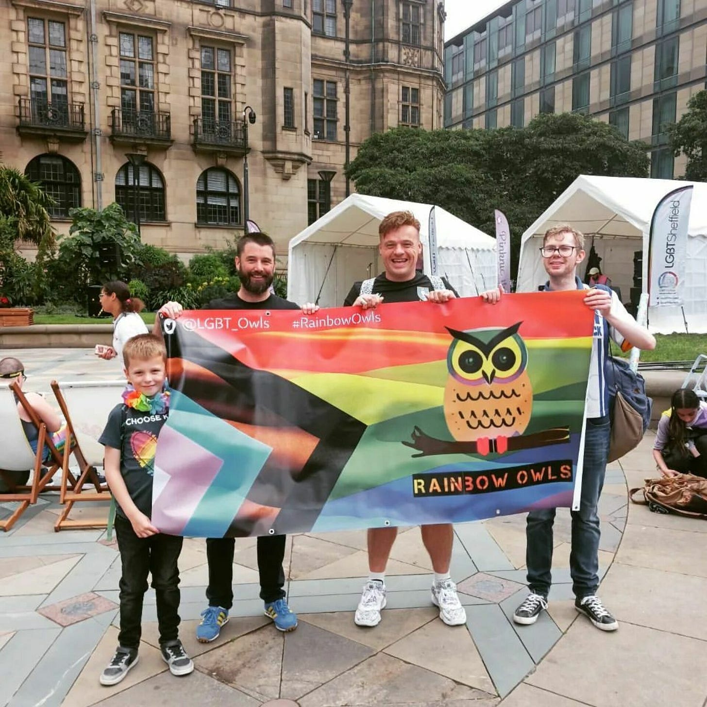 Three men and a boy hold up a Pride Progress flag with the rainbow owls logo printed on it.