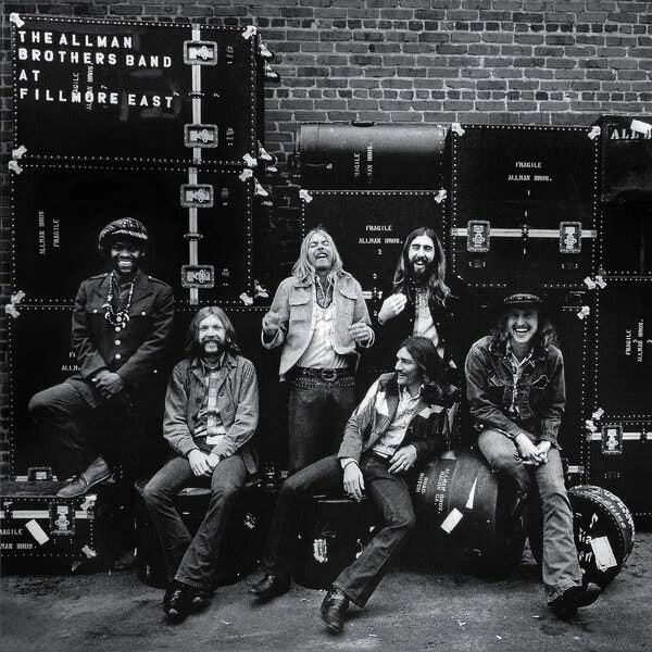 A black-and-white album cover, with a photograph of six casually dressed and longhaired musician leaning on and against equipment cases and the words “THE ALLMAN BROTHERS AT FILLMORE WEST,” in small white uppercase letters, in the upper left-hand corner.