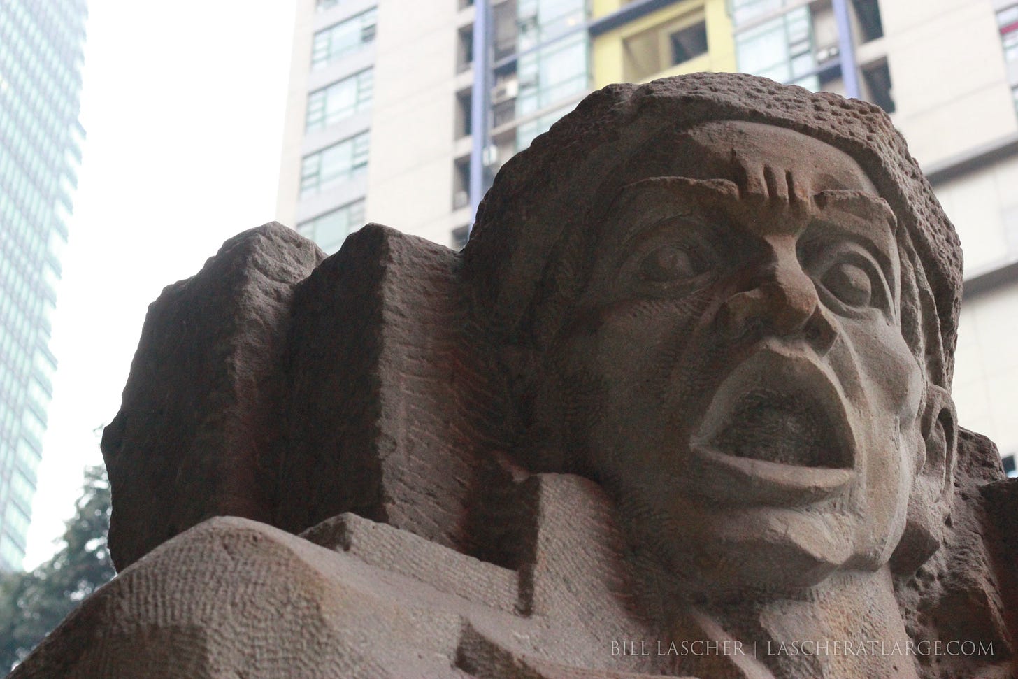 A carved stone face half in shadow carries a horrified, open-mouthed expression. The features are angular and stark. The face fill's the image's foreground while the windows of two large modern apartment towers are visible in the background.