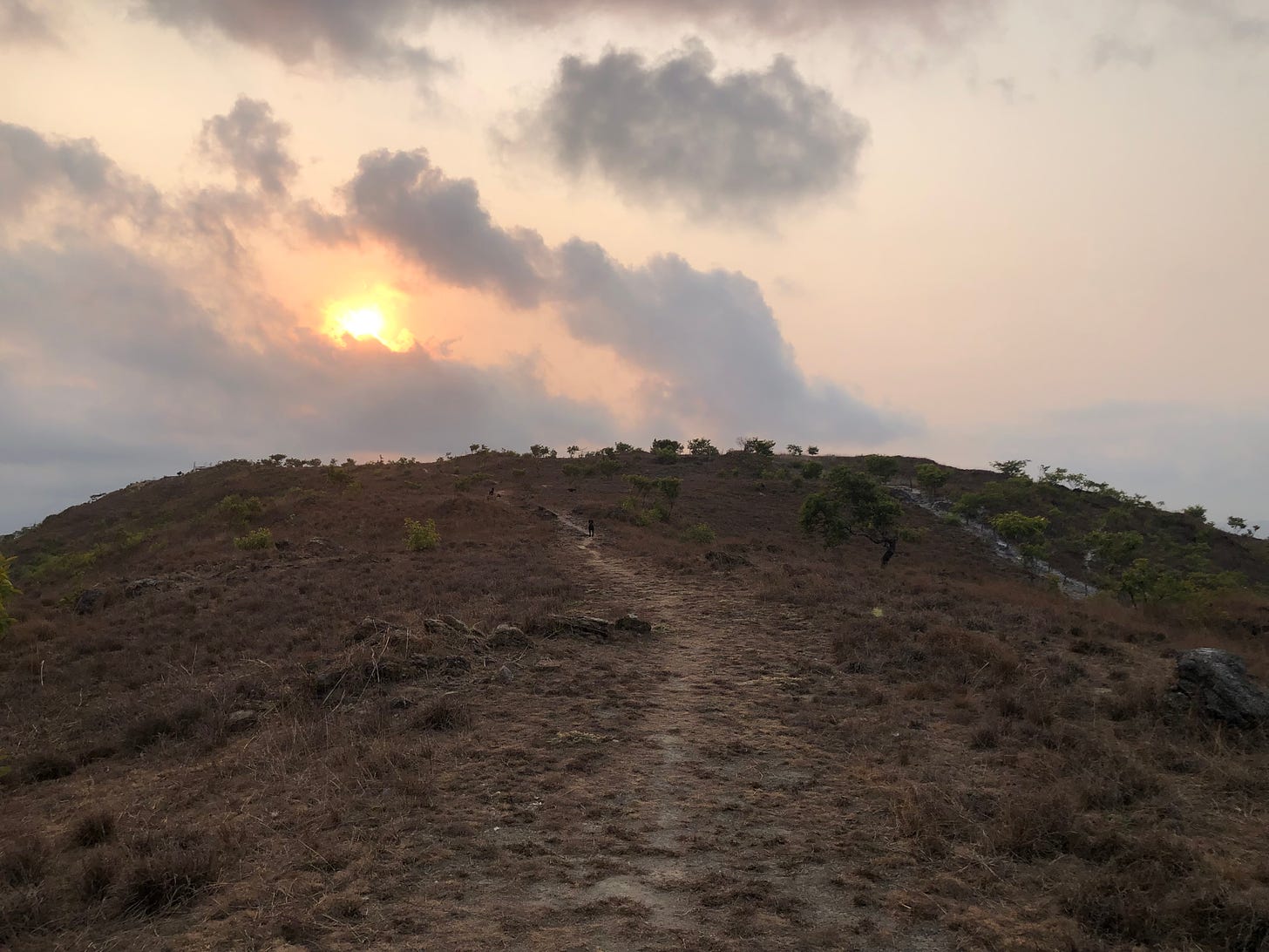 A hill covered in dead grass with a faded path down the middle, a sunset and runny clouds in the background.