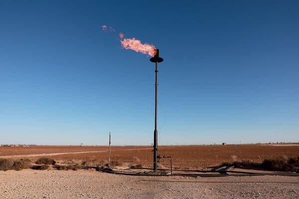 An orange flame shoots out to the left from the top of a tall vertical pipe that rises from a rust-colored desert landscape.