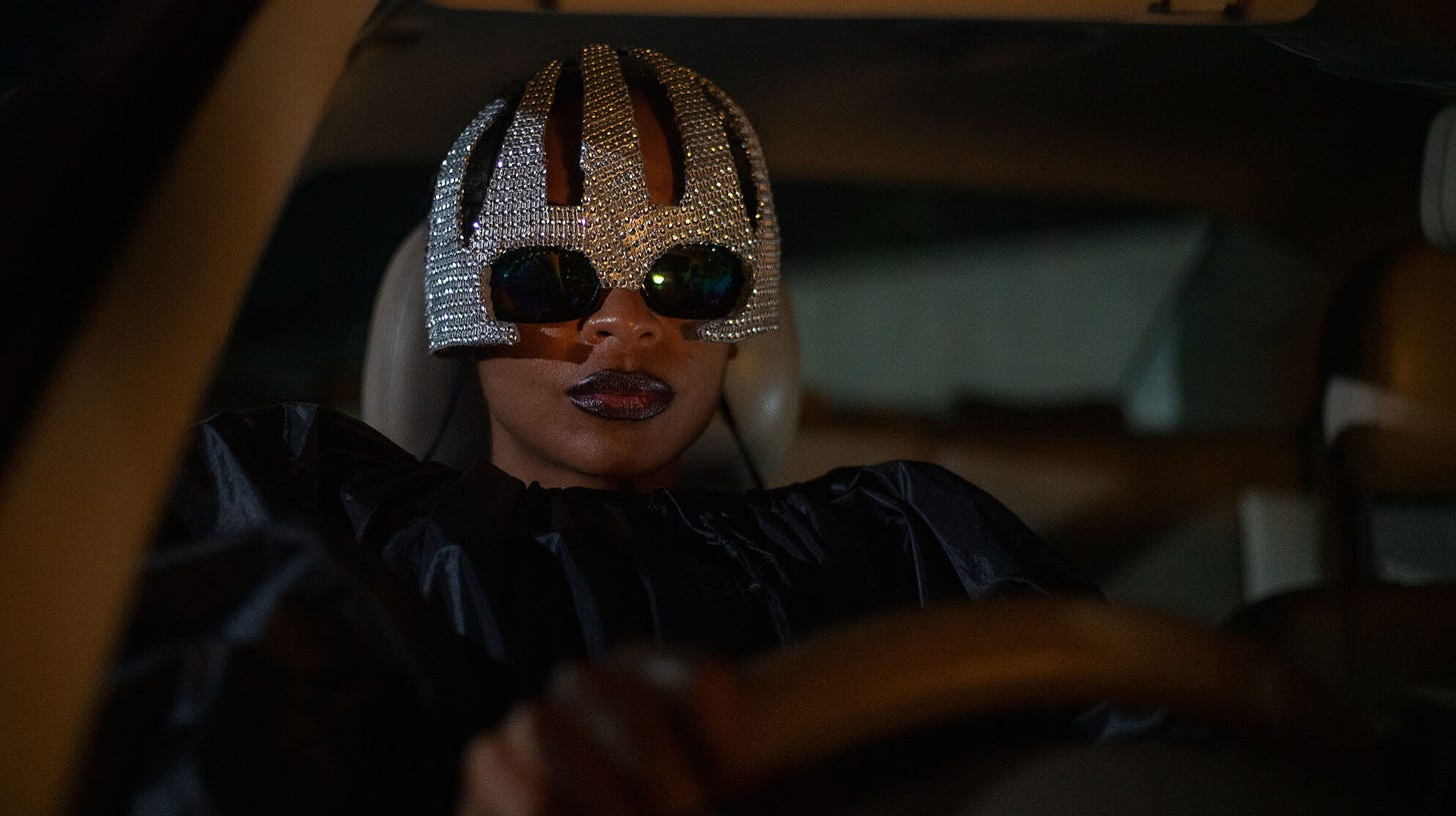 Shula (Susan Chardy) dressed as Missy Elliott in a black trashbag and a bejeweled helmet and sunglasses in "On Becoming a Guinea Fowl".