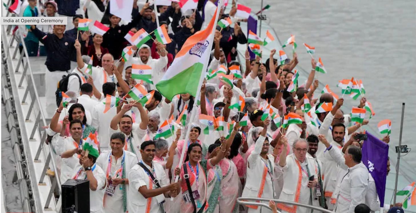 Indian athletes on their boat, wearing white jackets, waving small flags.