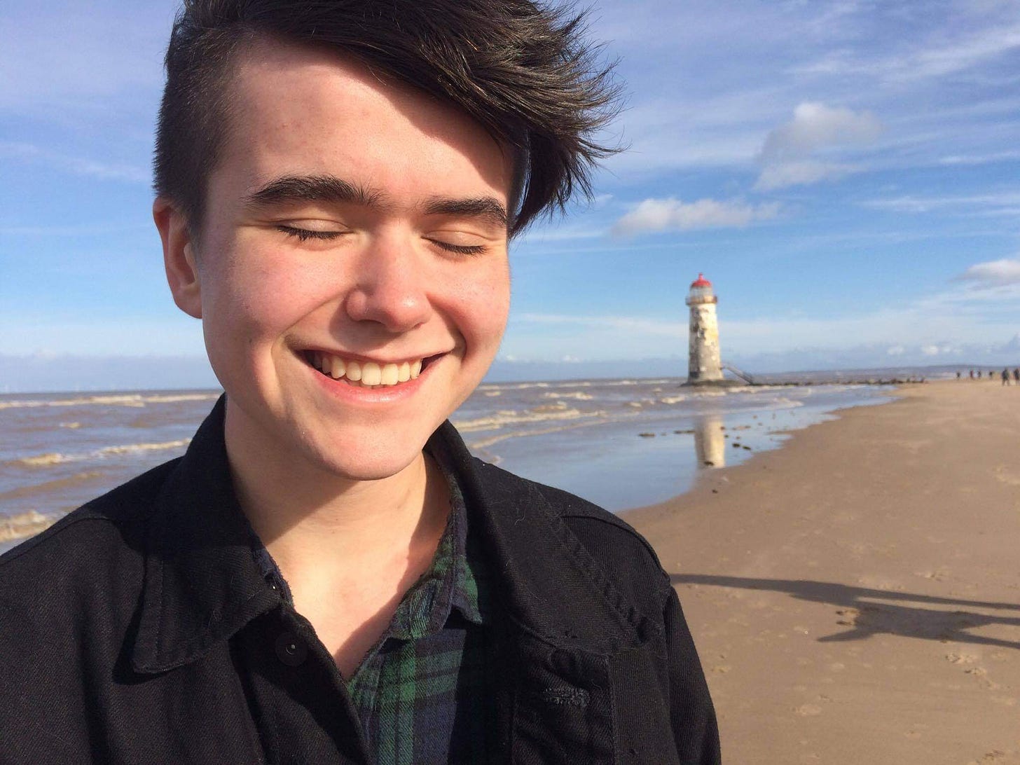 17 year old eliott on the beach, eyes closed and smiling. it is sunny and cold, and there is a battered lighthouse on the horizon