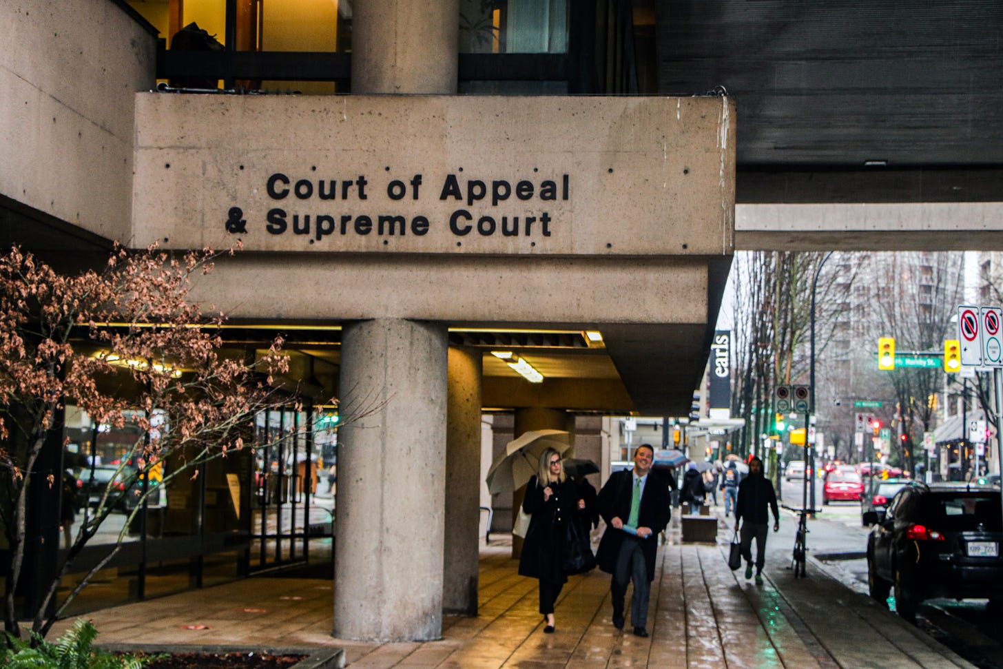 the entrance to the supreme court and court of appeal of BC in downtown vancouver on a rainy day, with people walking about in front of the entrance and beyond bearing umbrellas