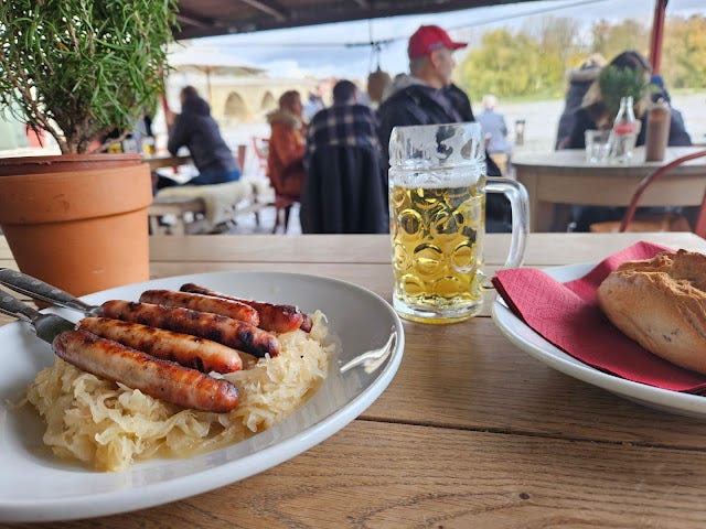 A plate of small sausages and sauerkraut and a small glass of beer, with the famous stone bridge across the Danube in the background