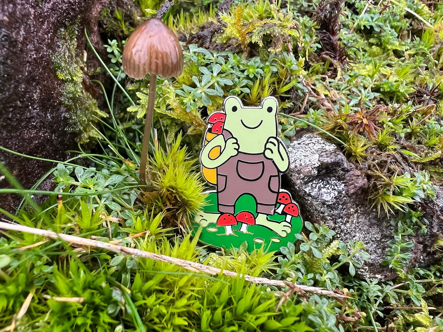 An enamel pin of a foraging frog in dungarees standing among red mushrooms