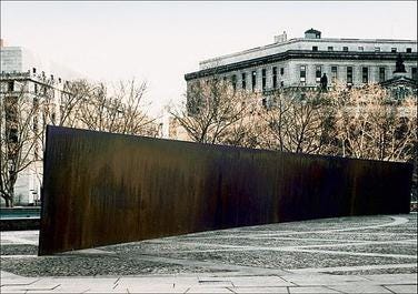 A rust-covered curved wall in a public square with buildings and trees in the background.
