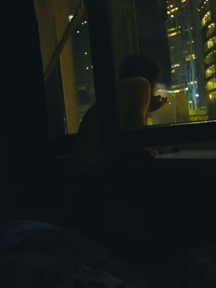 This may contain: a person standing in front of a window looking out at the night cityscape