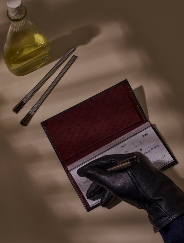 A gloved hand writes a check. Two brushes and a bottle of liquid sit on the table nearby.