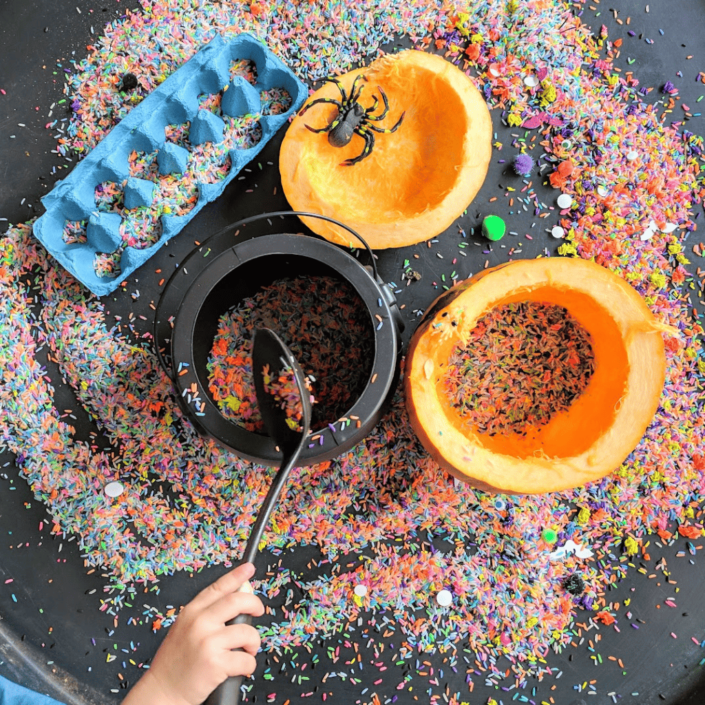 an overhead view of a pumpkin sensory play activity on a tuff spot tray featuring a hollowed out pumpkin, a plastic cauldron. Also visible is a toddlers hand as she scoops sensory rice into the pumpkin 