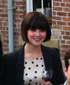 White woman with dark hair and bangs smiling at the camera. She's holding a glass of red wine and wearing a corporate black blazer and a spotty top