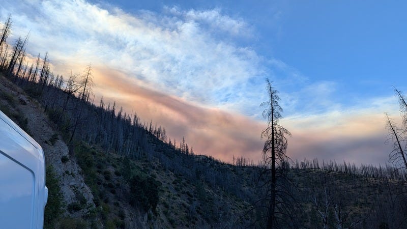 A photo of a hillside in shadow, blue sky above, a wall of smoke visible over the hill