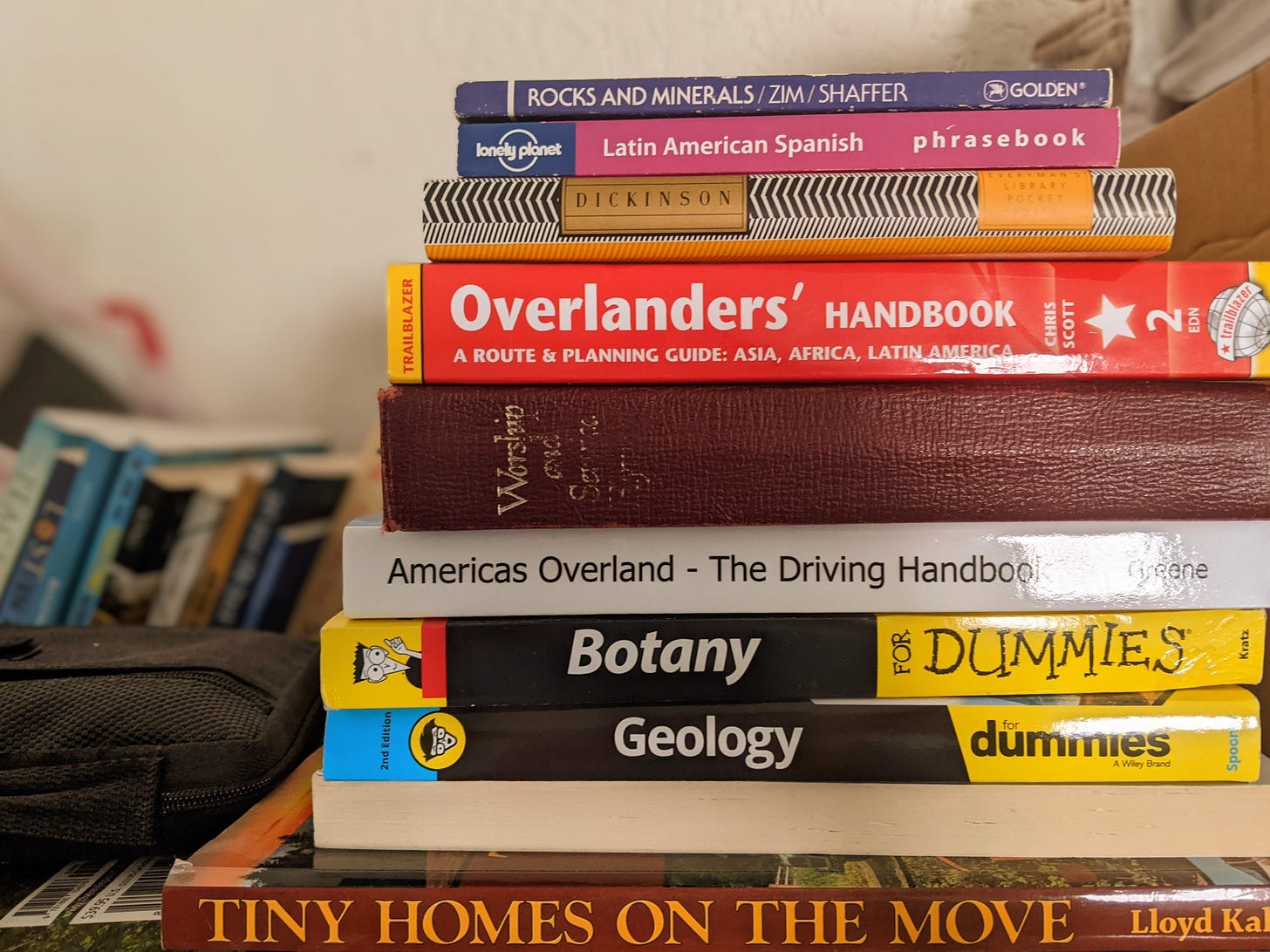 Stack of books in front of a row of more books. The books in the foreground are various sizes and on various topics related to travel and poetry.