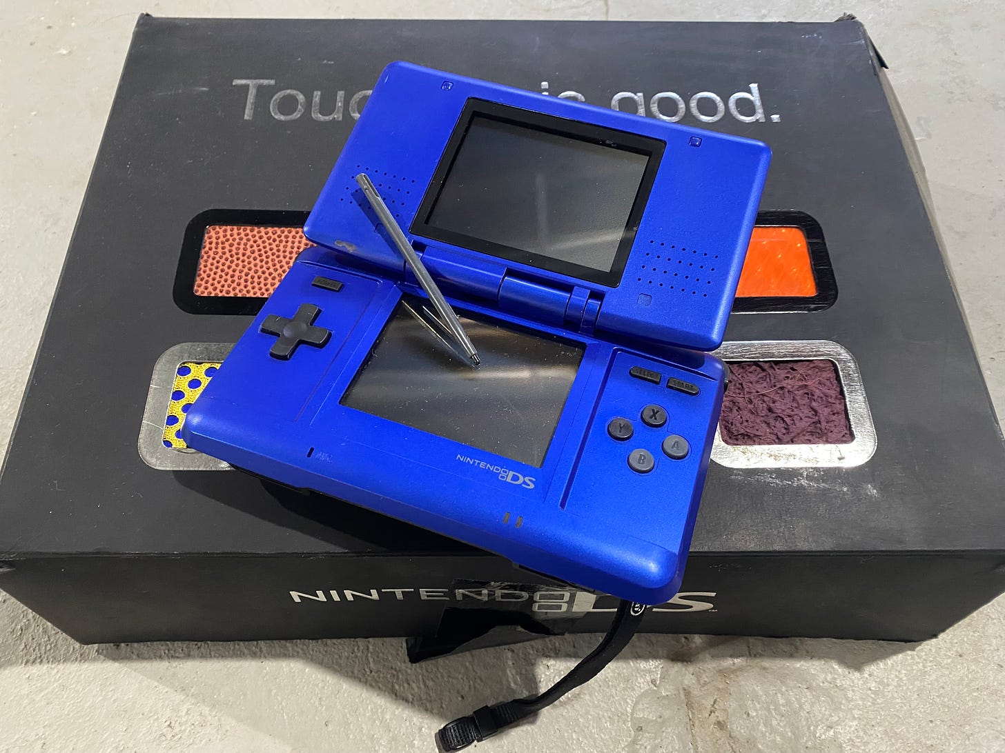 Photo of a blue Nintendo DS on top of a black box that bears the phrase "Touching is Good" and includes some dsquare panels of different rough textures.