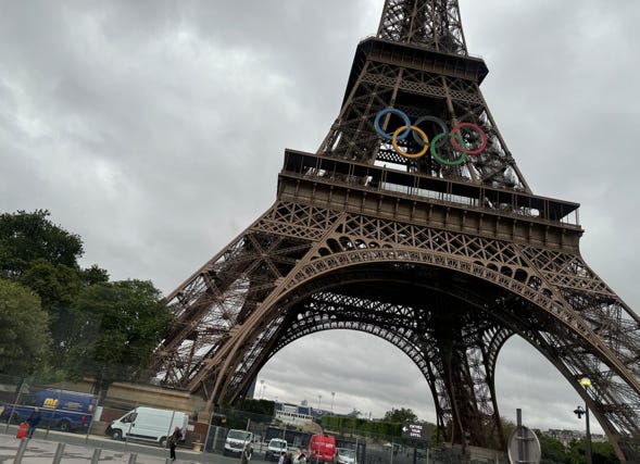 A large metal tower with rings on top with Eiffel Tower in the background

Description automatically generated