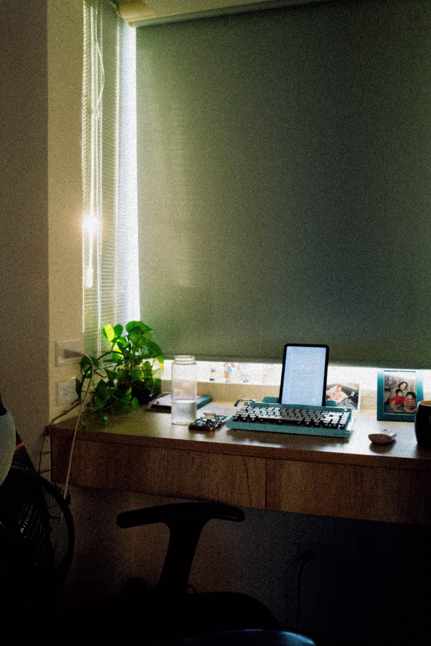 A desk by a window holds a tablet on a teal typewriter, a jar of water, a small plant, a framed family photo, and a black coffee mug. Sunlight filters through partially closed blinds.