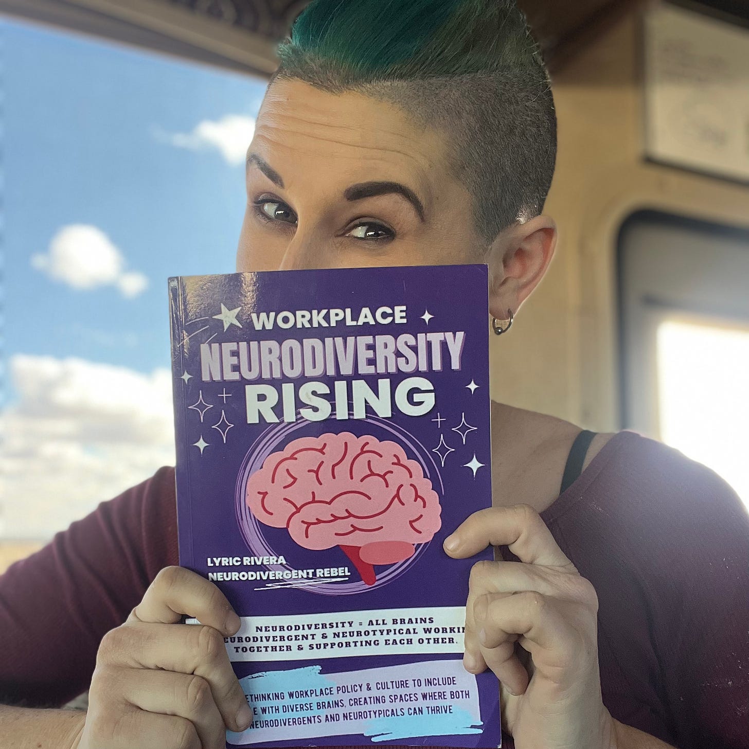 Photo of Lyric Rivera, a fair skinned nonbinary human with short green hair and shaved sides, holding up a purple book with a pink brain on the cover titled "Workplace NeuroDiversity Rising