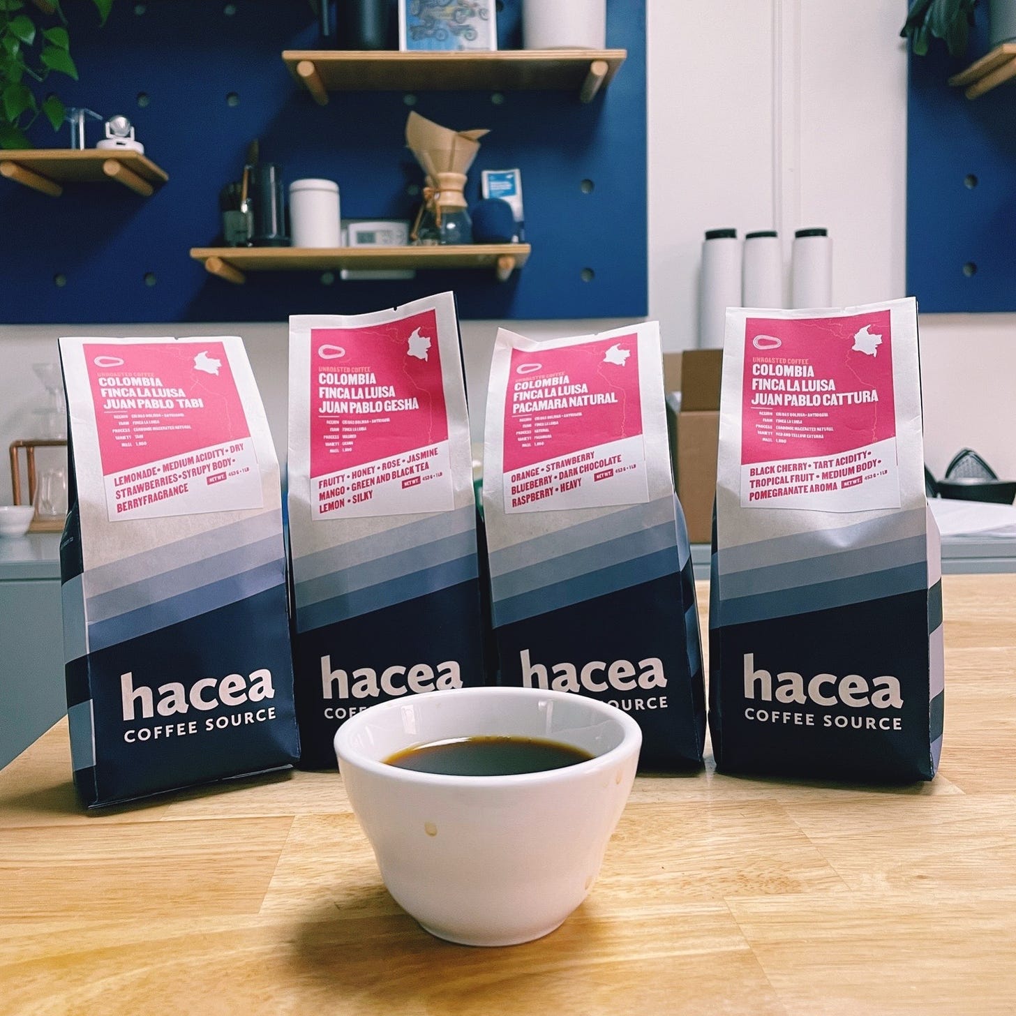 Four blue and white coffee bags with red labels at the top showcase Colombia coffees from Hacea Coffee Source. A coffee in a white cupping bowl sits in front of the packages.