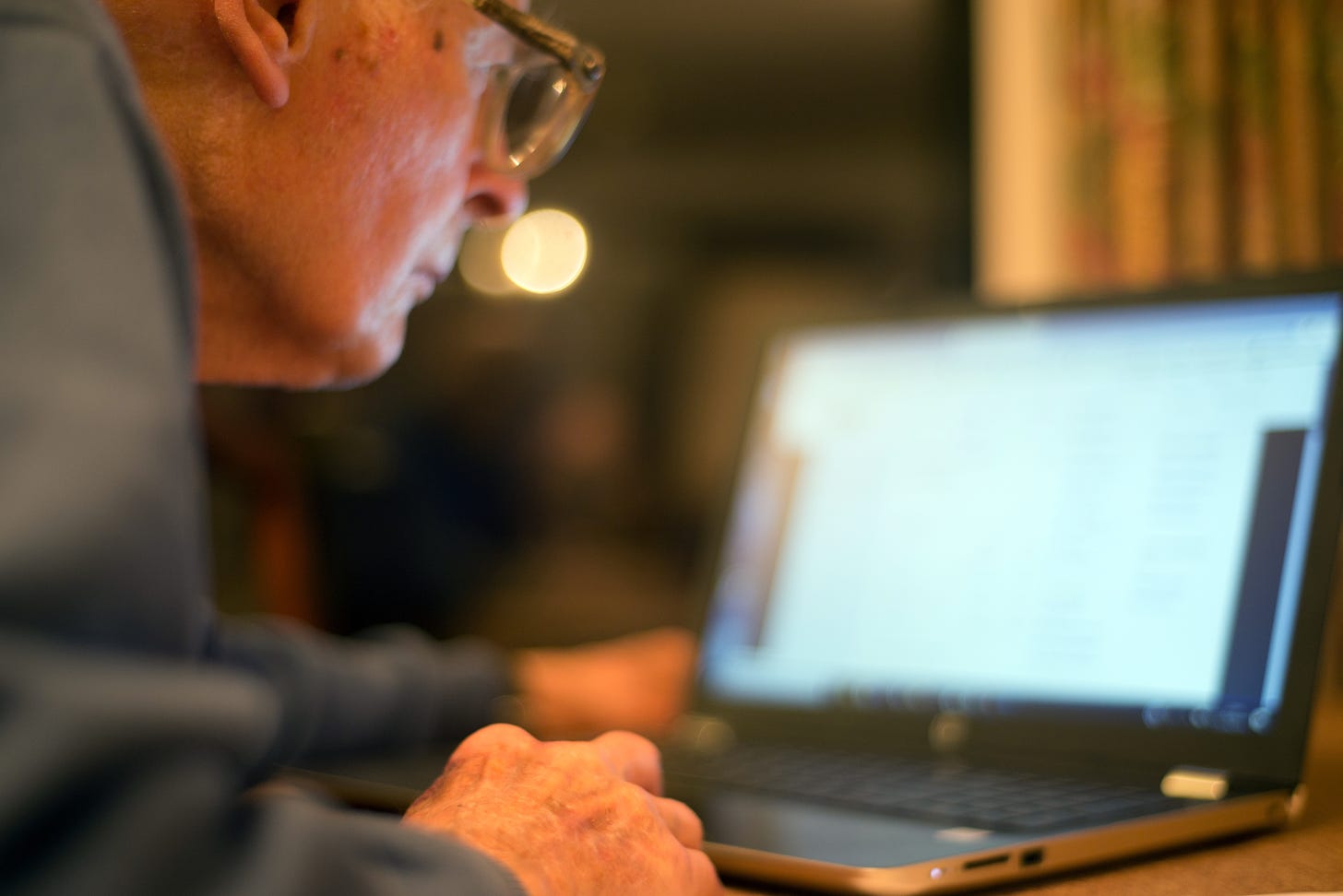 An elderly man wearing glasses leans forward and looks at a laptop.