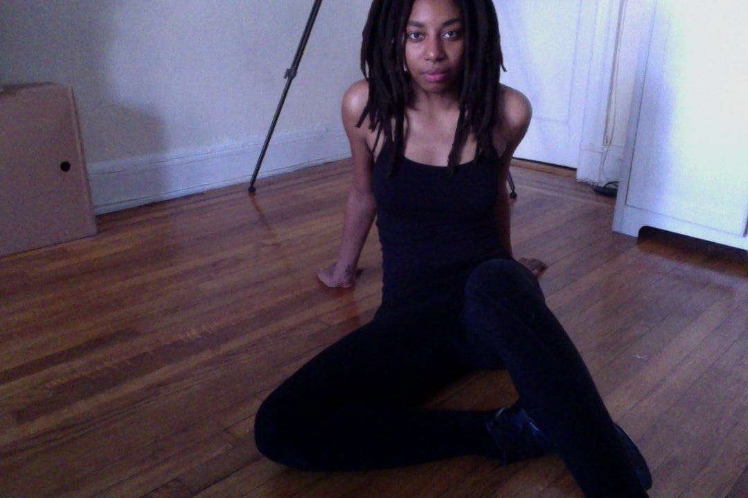 Young black woman with dreadlocks sits on floor wearing all black bodysuit and boots while staring straight into the camera