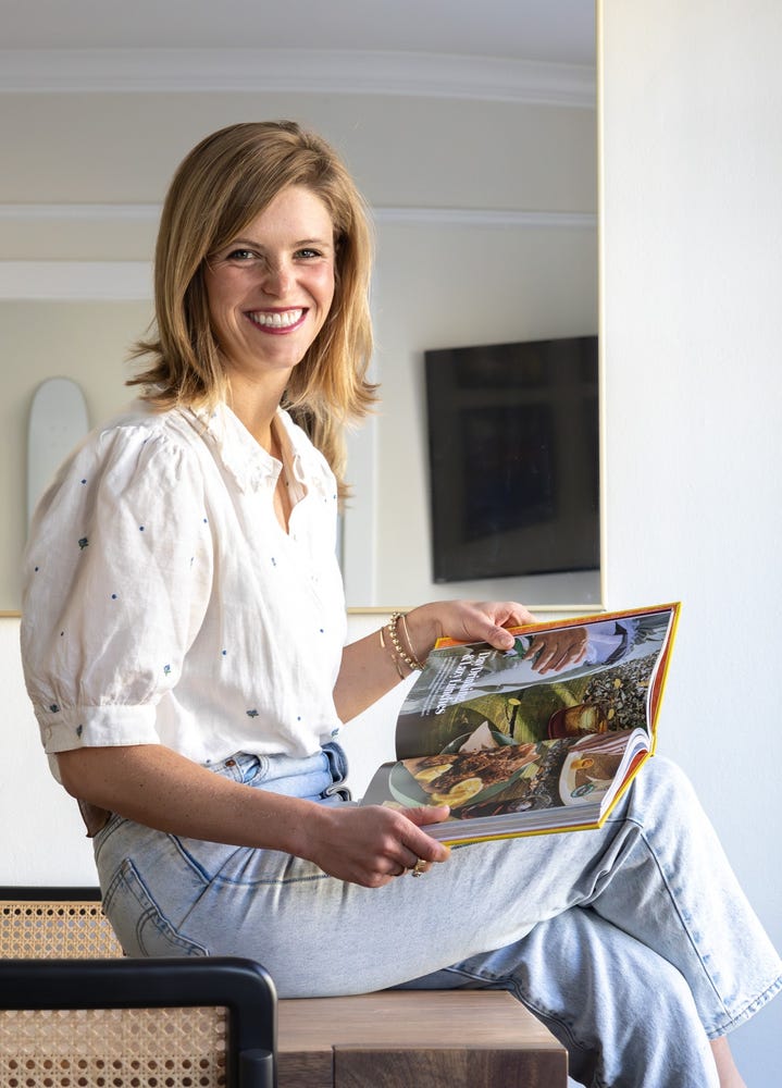 Anja sitting on a table legs crossed holding a cookbook smiling at the camera