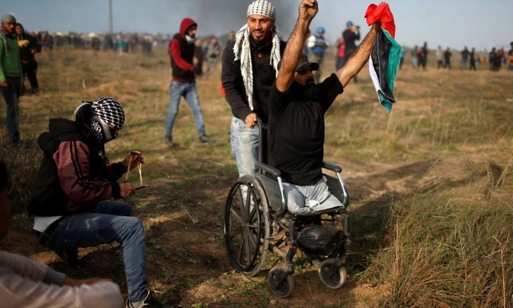 Photograph of Ibrahim Abu Thuraya in Gaza. He sits in a wheelchair hands raised with a palastinian flag grasped in one fist. He is in a field with a mass of people blurry in the background. 
