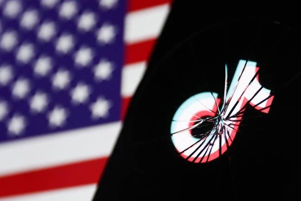 TikTok logo displayed on a phone screen is seen through the broken glass with American flag displayed on a screen in the background in this...