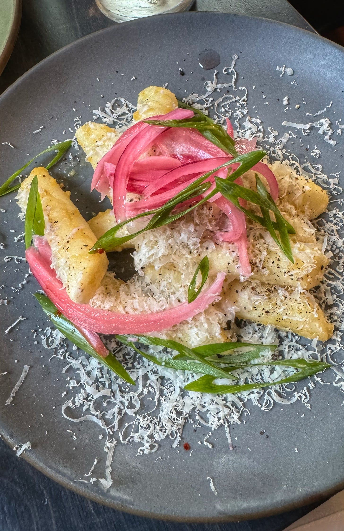 A plate of cacio e pepe tteokbokki, rice cakes showered in shaved cheese, slivers of pickled onions and thinly sliced green onion