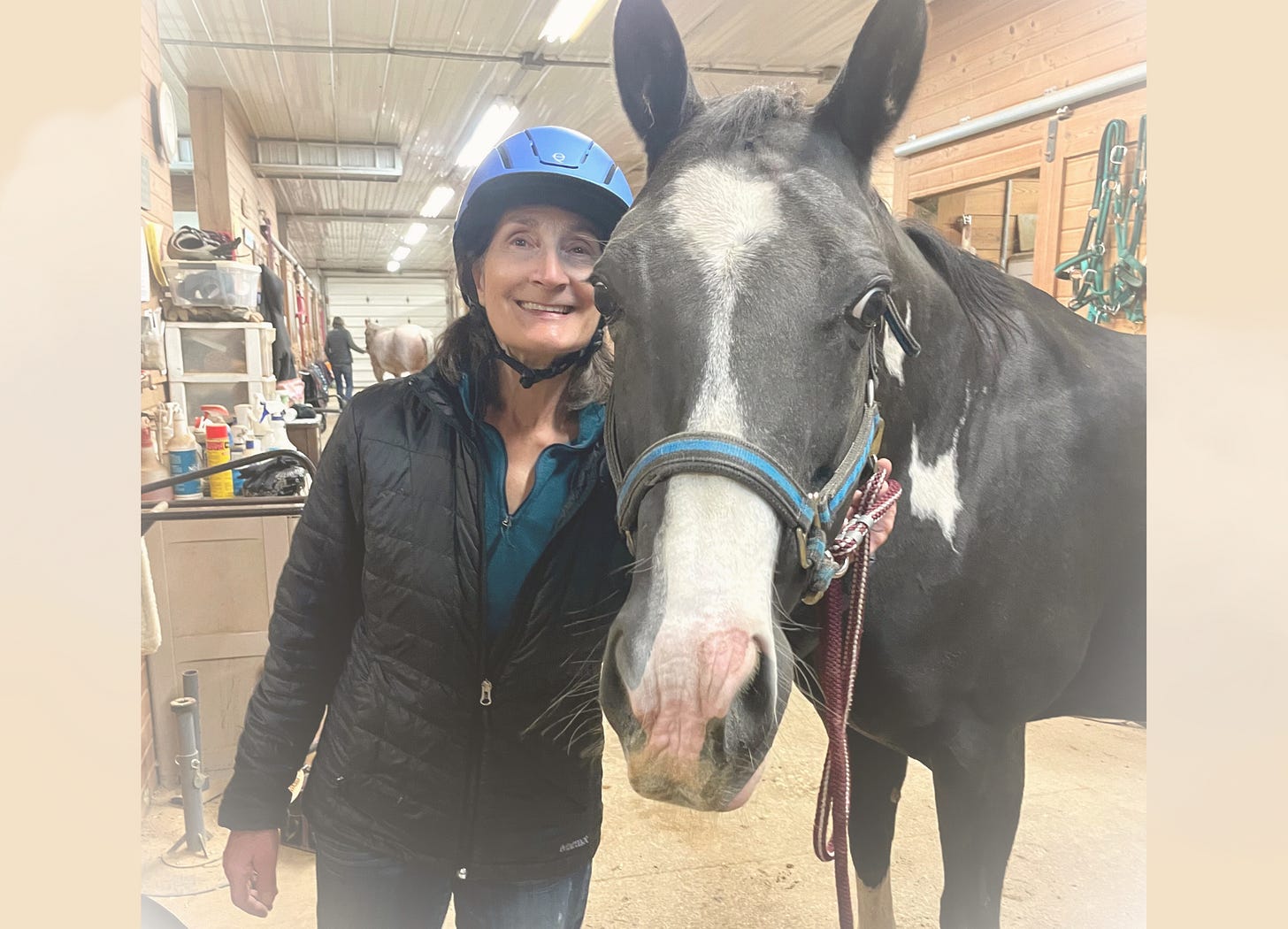 woman in black coat and blue riding helmet next to black horse with white nose