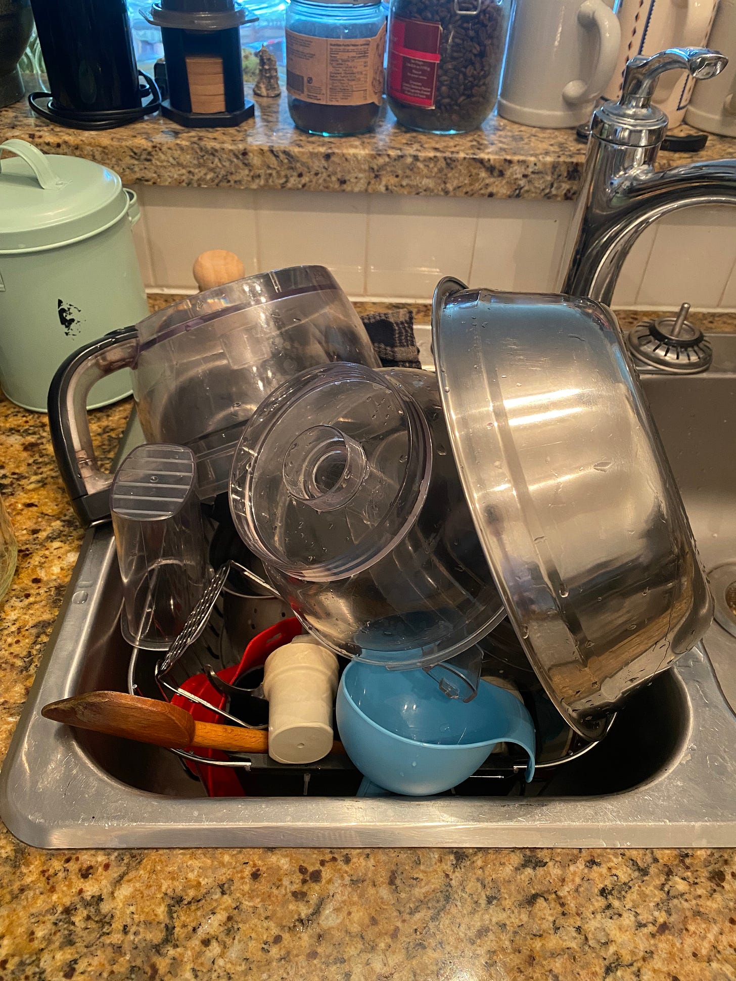 A sink full of dishes drying in the rack: a large bowl, a food processor, a masher, a couple of funnels, and a wooden spoon are visible.