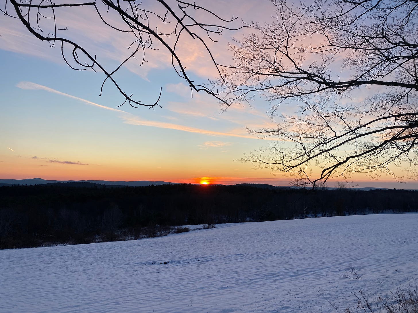 The golden sun rising behind dark hills in front of a snowy field.