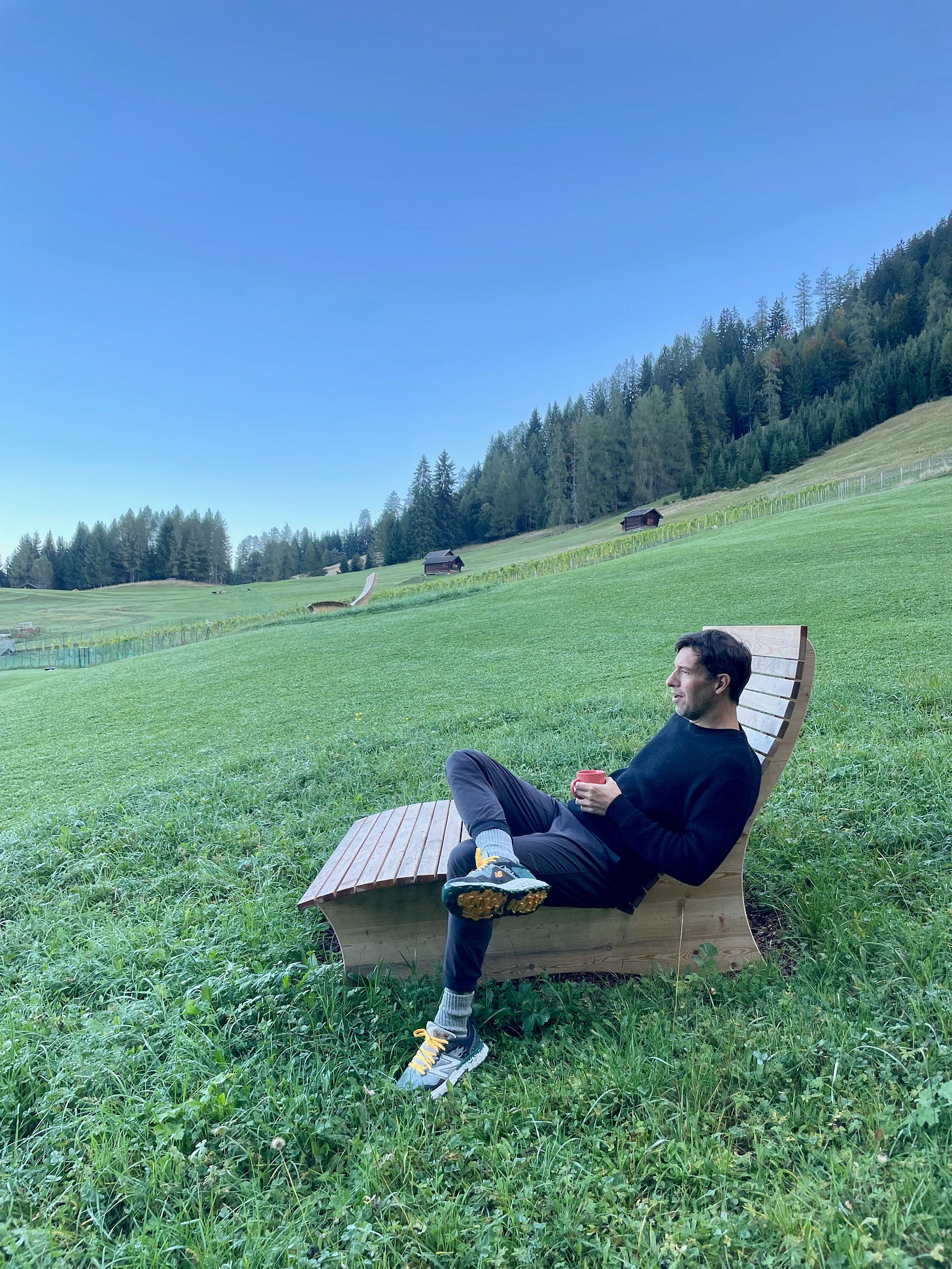 Chris Z sitting on a bench on a green field with a cup of coffee in his hands