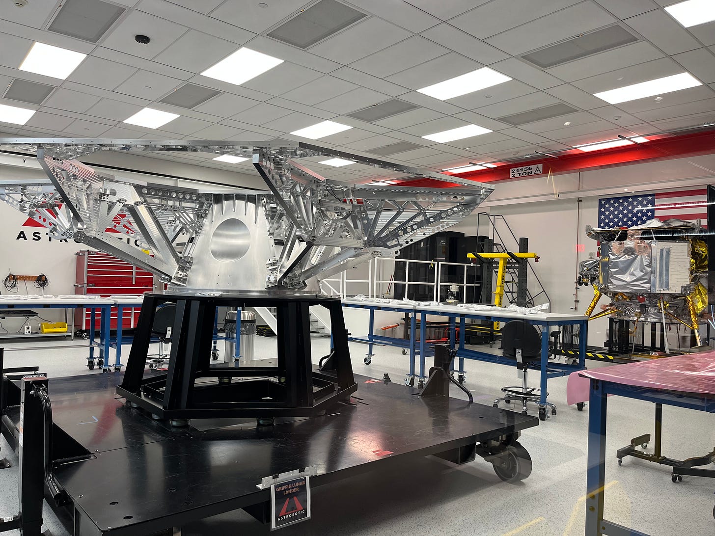 A photo of a smallish cleanroom. In the foreground is the bare metal skeleton of the Griffin lander, shaped roughly like a large satellite dish. In the background is the smaller but more complete Peregrine lander, a squat shiny silver box with spindly legs covered in gold foil.