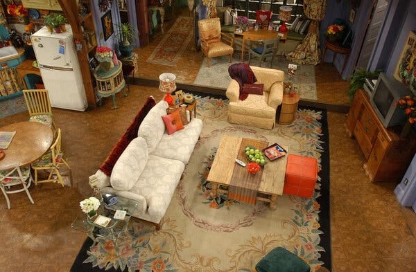 A view onto the set of Monica’s apartment in “Friends.” The walls are purple. In the center, a white sofa, a yellow armchair, a square wooden table with a bowl of green apples, on a floral carpet.