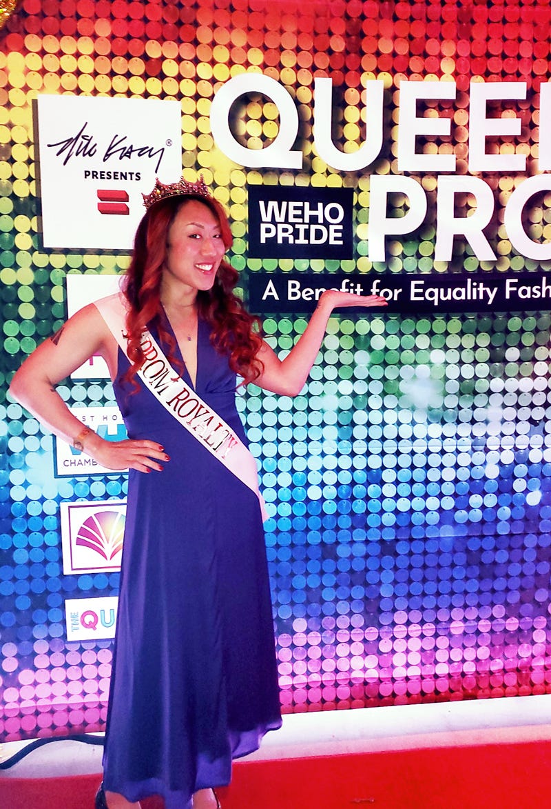 Alt text: An Asian-american Trans person wearing a purple dress, crown, and sash that reads “Prom Royal” stands in front of a rainbow banner for Queer Prom, an event put on for West Hollywood Pride that benefits Equality Fashion Week.