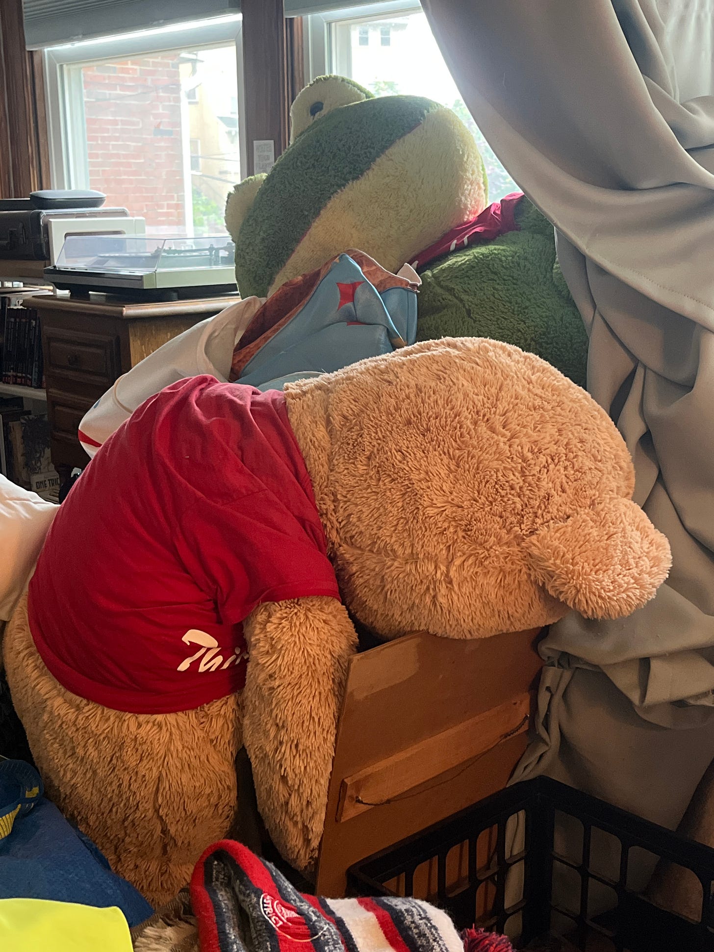 A giant stuffed teddy bear in a red tee shirt lies collapsed over a giant stuffed green frog. A giant stuffed teddy bear in a red tee shirt lies collapsed over a giant stuffed green frog.