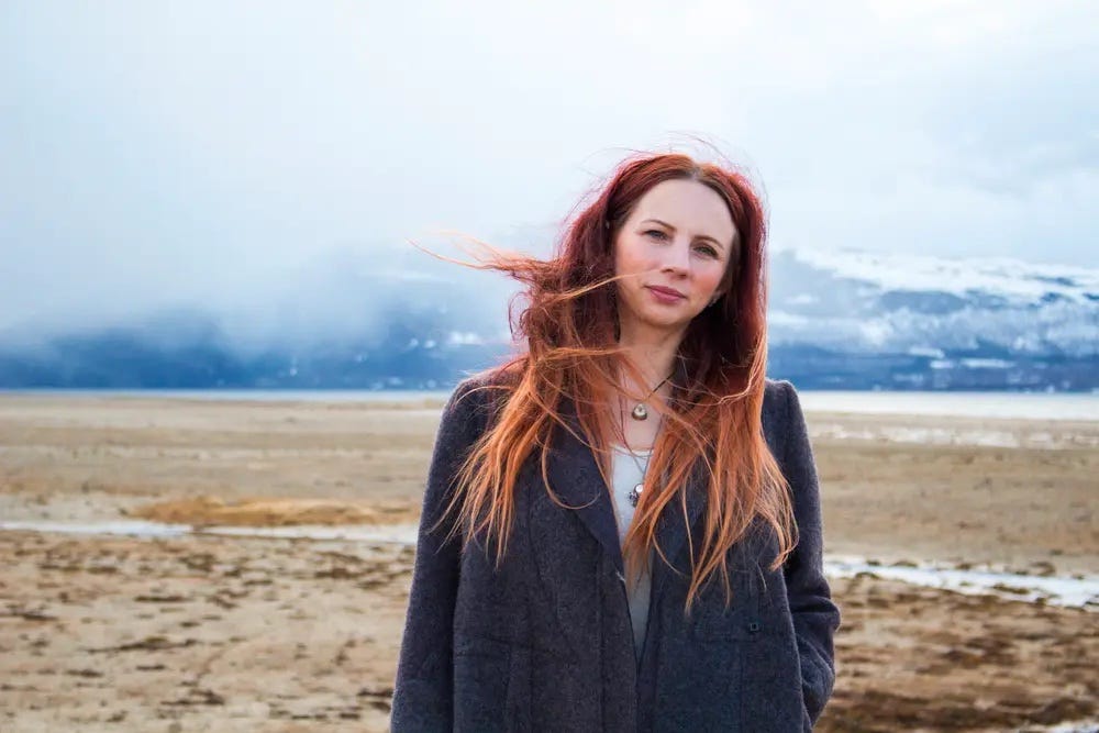 A photo of a white woman with red hair blowing in the wind. She stands in front of a snow capped mountain range. she wears a blue sweater and it is snowing