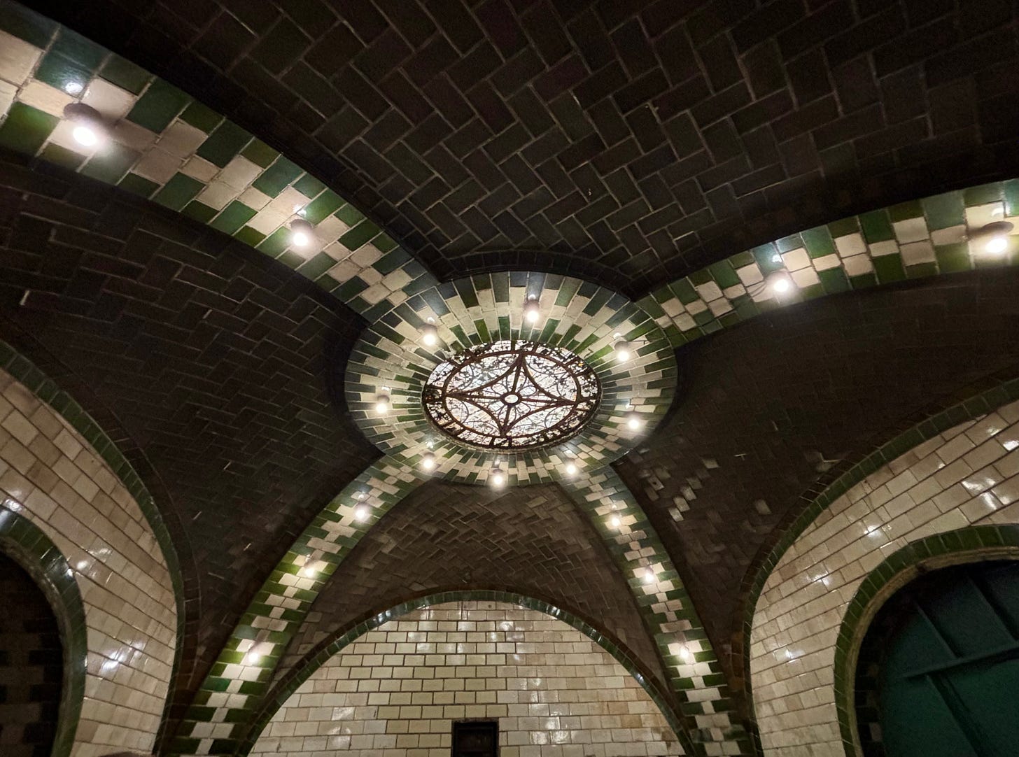 The high ceiling in the entrance of the old station. Green and white tiled arches cut across dark interlocking tiles. In the center is a decorate circular abstract pattern.