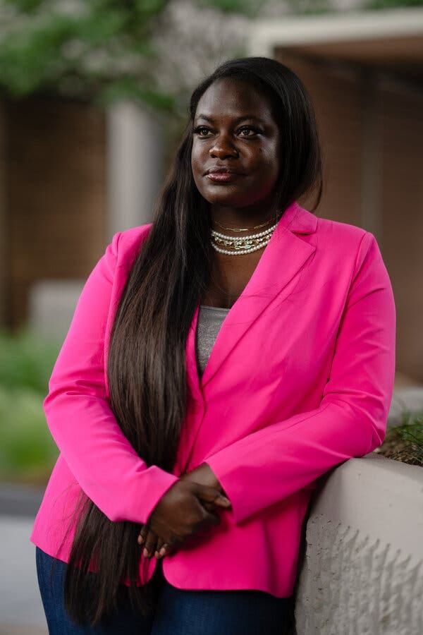 A woman in a bright pink jacket with long hair stands with her hands folded, looking off into the distance.