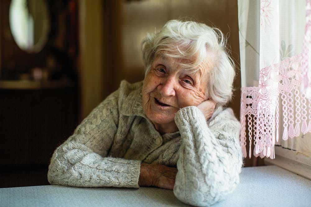 What’s too old? Elder woman smiling at the camera while sitting next to a window.